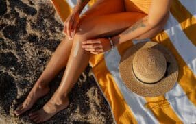 Women's legs on the beach on a striped towel and sun hat placed on the towel.